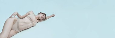 Pankart. Bej iç çamaşırlı manken yan yatar, kolları açılır, yumuşak ışık cilt üzerinde yumuşak gölgeler bırakır, açık mavi arka planda fotokopi alanı ile birlikte. Modern güzellik kavramı, sakinlik, özgüven.