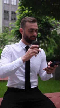 Ofis avlusunda oturan resmi kıyafetli bir iş adamı elinde kahve fincanı ve sakin bir şekilde telefonu kaydırıyor. Modern rutin kavramı, iş molası, bağlı yaşam tarzı.