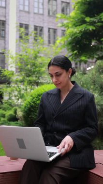 Ofis binasının yanında şık bir kadın yönetici oturuyor, dizüstü bilgisayarla çalışıyor, elleri sakin ve konsantre bir şekilde daktilo ediyor. Mobil ofis kavramı, kurumsal rutin, teknoloji destekli kariyer.