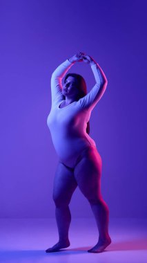Mor neon ışıkta koyu bir görsel kontrastla sergilenen, kendine güvenen ve güçlendiren iri yarı bir kadının tam görüntüsü. Beden pozitifliği ve güçlendirme kavramı.