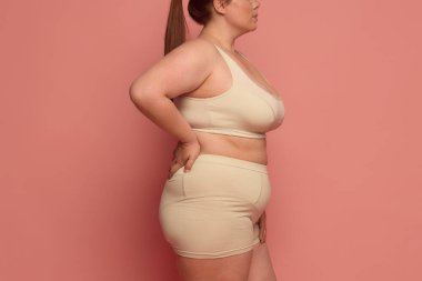 Bir kadın, pembe arka planda rahat bej iç çamaşırlarıyla doğal bedenini kucaklıyor. Beden pozitifliği ve kendini sevme kavramı.