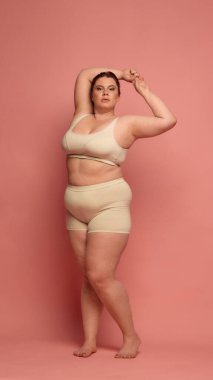 Kendine güvenen bir kadın pozitifliği kucaklar, pembe arka planda rahat nötr iç çamaşırı giyer. Kendini kabul etme ve güçlendirme kavramı.