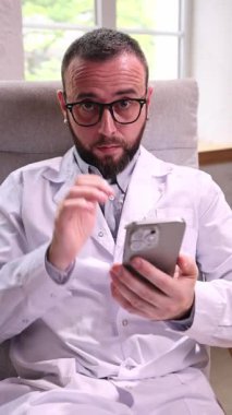 Laboratuvar önlüklü erkek doktor rahat bir şekilde ofis koltuğunda oturuyor, akıllı telefona kayıyor, kısaca yukarı bakıyor ve ekranda yakınlaşıyor, modern tıptaki dijital çoklu görevi sembolize ediyor.