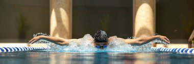 Bir yüzücü, kapalı havuzda kelebek vuruşunu gerçekleştirir. Kararlılık ve odaklanma gösterir. Atletik azim ve rekabetçi sporlarda kendini adama kavramı.