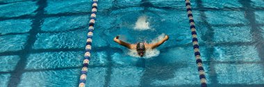 Genç bir erkek yüzücü, yüzme havuzu şeridinde güçlü bir kelebek vuruşu yapıyor. Su sporlarında atletik cesaret ve rekabetçi ruh kavramı.