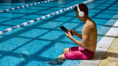 Kulaklıklı beyaz bir adam yüzme havuzunun kenarında otururken tablet kullanıyor. Yaz boyunca rahatlama ve dijital rahatlama kavramı.