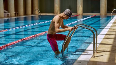 Beyaz bir erkek yüzücü kapalı havuzdan yüzme şapkası ve gözlüğü takarak çıkıyor. Rekabetçi yüzmede kendini adama ve azim kavramı.