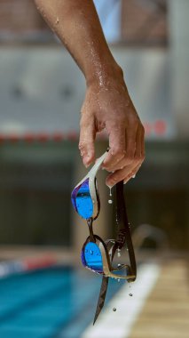 Bir el ıslak yüzme gözlüklerini havuz kenarında tutar, su damlacıkları lenslerden damlar. Ferahlatıcı bir yüzmeye hazır olma kavramı.