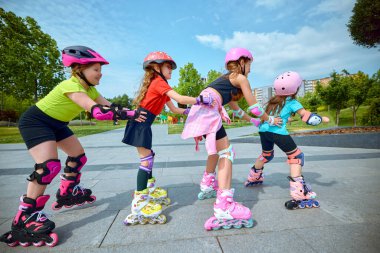 Küçük çocuklar açık, güneşli gökyüzünün altındaki bir parkta grup halinde paten kaymayı, açık hava aktiviteleri ve çocuklar için fiziksel uygunluk fikrini somutlaştırmayı seviyorlar..