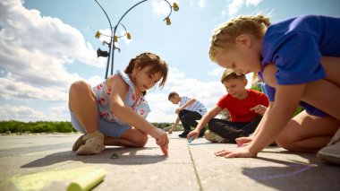 Çocuklar güneşli bir gökyüzünün altındaki kaldırımda renkli tebeşir çizimleri yaparlar. Yaratıcılık ve mutluluk gösterirler. Gençler için dışarıda sanat faaliyetleri fikri.
