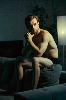 Ciddi bir ifadeyle siyah iç çamaşırıyla kanepede oturan dalgın erkek manken. Düşünceli ruh hali, ev modası, portre çalışmaları ve kişisel sağlık kavramı..