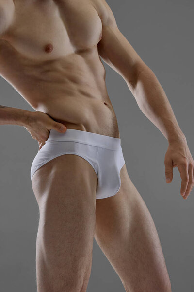 Атлетик уверенно позирует в белых трусах на сером фоне. Концепция ориентированной на тело рекламы, мужской физической формы и фитнес-идентичности