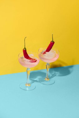 İki kokteyl bardağı, içinde donmuş kırmızı biber olan sarı mavi yüzeydeki buz küpleri. Baharatlı içecekler, yaratıcı karışım, gerçeküstü pop sanat içeceği reklamcılığı ve pazarlama kullanımı kavramı.