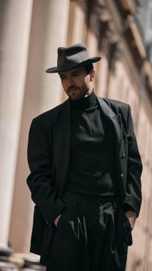 Siyah takım elbiseli ve fötr şapkalı bir adam arka planda taş sütunlarla dışarıda poz veriyor. Hikaye anlatımı tabanlı sanat alanları kavramı, zaman tabanlı medya sergileri