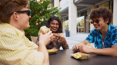 Dışarıdaki masada hamburger ve patates yiyen üç genç arkadaş. Sokak yemekleri reklamı, günlük sosyal anlar, gençlik yaşam tarzı tanıtımı, dağıtım platformu görselleri..