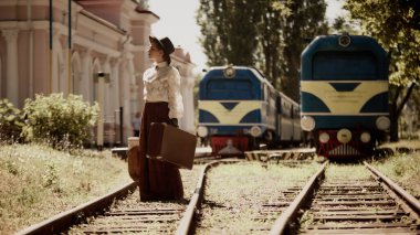 Antika elbiseli bir kadın iki mavi trenin yanında bavuluyla demiryolunda duruyor. Tarihsel seyahat kavramı, dönem kurgu hikayesi, geçiş kültürü, romantik anlatı.