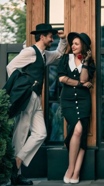 Şapka takan ve siyah elbiseli kadına gülümseyen bir adam ahşap duvarın yanında duruyor. Romantik roman kapağı kavramı, tiyatro posteri, insan ilişkileri illüstrasyonu..