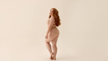 Tarafsız arka planda çıplak iç çamaşırlarıyla poz veren kendinden emin, iri yarı bir kadın. Anlamlı hareket kavramı, neşe ve kıvrımların kutlanması, iç çamaşırı için enerjik markalaşma..