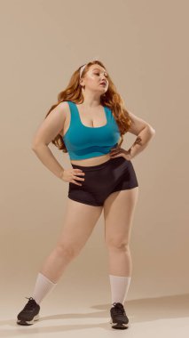 Kendine güvenen, spor elbiseli, elleri kalçasında bej arka planda duran bir kadın. Spor kıyafetleri anlayışı, kapsayıcı fitness markalaşması, tüm vücut tipleri için aktif yaşam tarzı anlayışı.