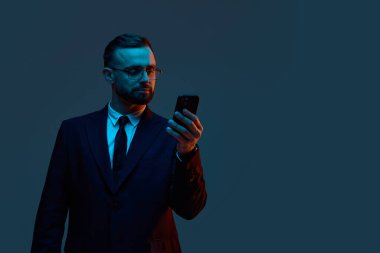 Takım elbiseli bir işadamı mavi ışık altında odaklanmış akıllı telefona bakar. Güvenli dijital ortamlarda mobil bankacılık, fintech arayüz testi ve iş iletişimi kavramı.