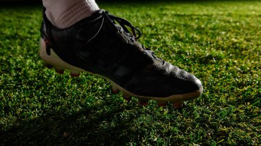 Sahne ışıkları altında çimlere bastıran futbol kramponlarının yakın çekimi. Futbol gerilimi, maç başlangıcı, erkek sporcu mevcudiyeti, bölge ile odaklanma ve fiziksel temas.