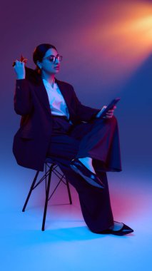 İş kadını akıllı telefonu tutarken neon ışığı altında saçlarıyla oynuyor. Mobil iş rutini kavramı, fintech liderlik ve rahat odaklı dijital yaşam tarzı.