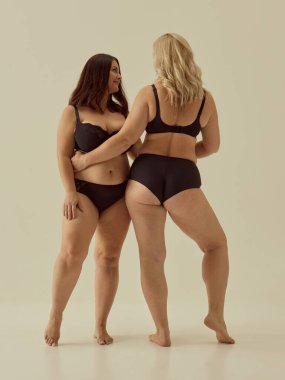 Siyah iç çamaşırlı iki kadın tarafsız arka planda vücut pozitifliği sergiliyor. Gerçek vücut tipleri için kadın iletişimi, neşeli hareket, kendini ifade etme ve moda reklamcılığı kavramı.