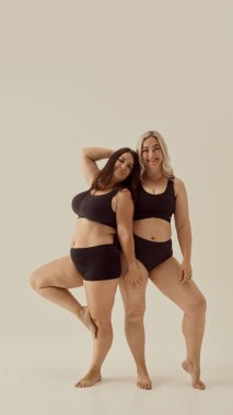 Tarafsız bir arkaplanda siyah iç çamaşırlı iki kendine güvenen kadın. Cesur enerji kavramı, kadınsı güç, büyük beden hareketleri ve dışavurumsal vücut sunumu..