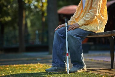 Güneş ışığında parktaki bankta otururken, elinde değnek tutan kör adamın yakın çekimi. Yardımcı araçlar, hareketlilik, kapsayıcı tasarım ve kentsel erişilebilirlik kavramı.