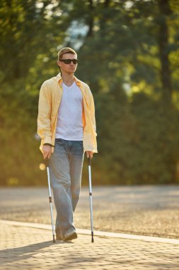 Asistan yürüyüş direklerini kullanarak parkın yolunda bağımsız yürüyen kör bir adam. Asistiv teknoloji, sosyal kapsama, bağımsız seyahat ve erişilebilir tasarım kavramı.