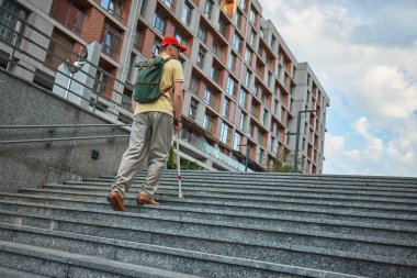 Kırmızı şapkalı kör adam beton merdivenlerden beyaz bastonla çıkıyor. Hareket kabiliyeti, kentsel erişim, bariyersiz tasarım, kişisel güç ve kapsayıcı altyapı kavramı.