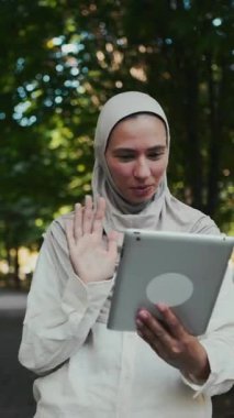 Açık renk tesettürlü, elinde tablet tutarken gülümseyen ve el sallayan bir kadın. İş iletişimi, bağlantı ve uzak toplantılar kavramı.