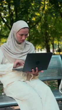 Bej tesettürlü ve açık renk elbiseli Müslüman bir kadın park bankında otururken dizüstü bilgisayarla yoğun bir şekilde çalışıyor. Uzaktan çalışma kavramı, doğadaki teknoloji, kadın odaklanma ve açık hava verimliliği..