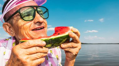Suyun kenarında bir dilim karpuz tutarken gülümseyen mutlu kıdemli adam. Neşeli yaşlanma, yaz tatili, sağlık, sağlıklı beslenme, seyahat eğlencesi ve kaygısız anlar kavramı..