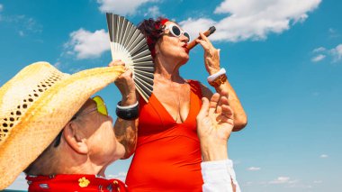 Kırmızı mayo giymiş, mavi gökyüzünün altında puro içen şık, yaşlı bir kadın. Aktif yaşlanma, kendine güven, boş zaman yolculuğu, yaz yaşam tarzı, refah, kişisel özgürlük kavramı.