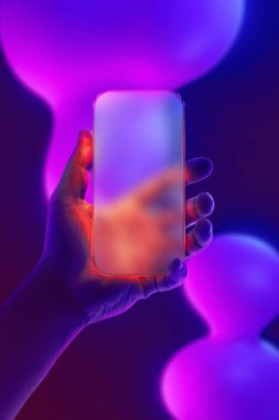 Sıvı cam tasarımı ve bulanık yansıması olan, mor, mavi ve pembe renkte neon sıvı şekillere ayarlanmış, elle kavrayan parlak bir akıllı telefon. Teknoloji, yenilik ve modern dijital estetik kavramı