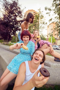 Arkadaşları renkli çörekler tutuyor ve yaz gezisi sırasında gülümsüyor. Arkadaşlık kavramı, lezzetli tatlılar, şehir eğlencesi, neşeli yaşam tarzı ve paylaşılan neşeli anlar..