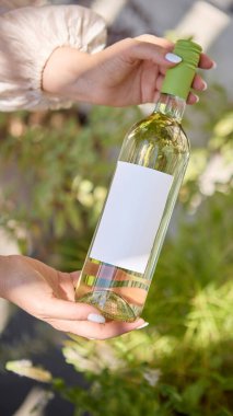 Boş etiketli beyaz şarap şişesi iki elle tutulmuş ve yeşillik var. Alkol turizmi, yaşam tarzı içeriği, reklam kampanyaları, menü tasarımı ve sosyal medya markalaşması kavramı.