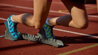 Sporcu, sakin bir odak ile mavi koşu ayakkabılarıyla başlangıç bloklarını hazırlıyor. Konsantrasyon kavramı, blok ayarlamasına başlama, atletik hazırlık ve rekabete hazırlık.
