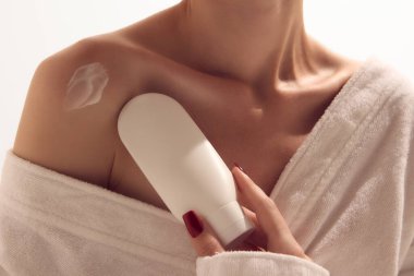 Bornozlu kadın, cilt bakımı ve güzellik markası için kozmetik şişeyle omuzuna losyon sürüyor. Kişisel bakım, kaplıca tedavisi, kozmetik reklamcılık ve kadın sağlığı yaşam tarzı kavramı..