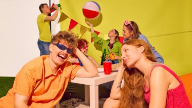 İçkiler ve plaj topuyla masada oturan bir grup gülümseyen genç arkadaş. Gençlik bağı, kaygısız yaşam tarzı, renkli kutlama, popüler kültür enerjisi ve neşeli hafta sonu kavramı..