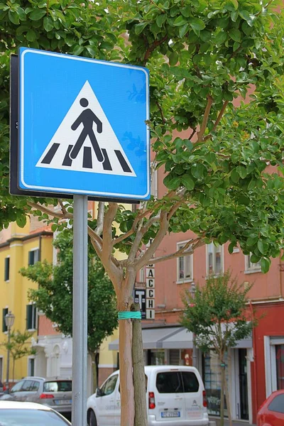 Yaya geçidi tabelası, mavi uyarı işareti, caddede kavşak işareti olan cadde, evler arasında ağaçlar.