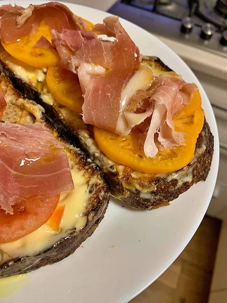toasted slices of bread, toasted slices, toast with melted cheese, slices of yellow tomatoes, raw Parma ham, snack on a plate