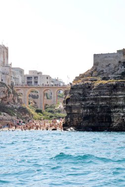  Kumsalda köprü, Polignano 'da körfez, İtalya' nın güneyinde basit beyaz binalar, kıyıda düz kayalar, kayalar arasında sahil, kayalık sahil.
