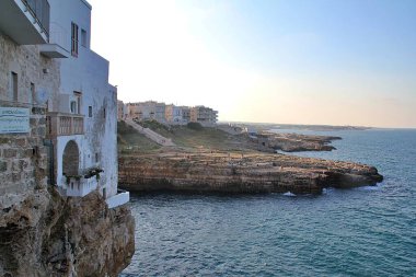 Deniz kıyısındaki büyük uçurum, deniz kenarındaki kayalar, deniz kıyısındaki kayaların üzerine inşa edilmiş binalar, Polignano a Mare, kayalık sahil, turkuaz su, denizde yıkanan insanlar.