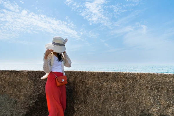 Woman wearing a white cardigan sweater and red pants is hiding behind hat with a blue sea and sky background