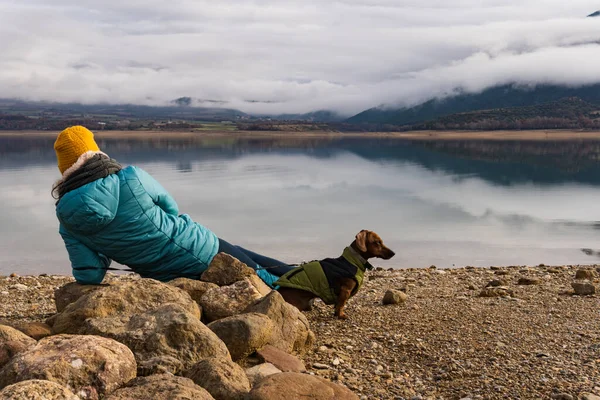 Rear view of unrecognizable woman resting on the rock with her dachshund teckel dog near to the lake where the mountains and clouds are reflected. they are wearing warm clothe