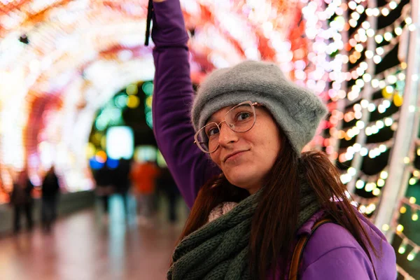 Smiling young woman on well decorated town square with christmas lights wearing glasses, hat and jacket