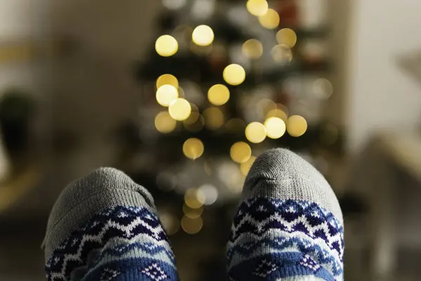 Close up of feet wearing warm Christmas socks with illuminated Christmas tree on the back ground