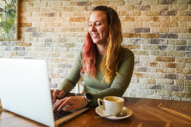 Kahvesinde dizüstü bilgisayar kullanan gülümseyen bir kadın..